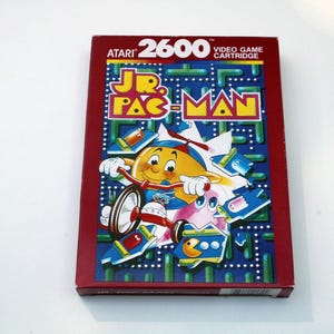May include: A rectangular Atari 2600 Jr. Pac-Man video game cartridge box with a deep red border. The front features a yellow character riding a tricycle, surrounded by blue maze walls and smaller game icons against a dark blue background.