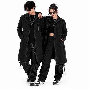 May include: Two individuals wearing matching black denim trench coats with silver button closures, vertical zipped chest pockets, and side waist straps. They are paired with black cargo trousers featuring side pockets and black and white trainers.