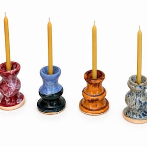 May include: Four ceramic candle holders with a rounded, tiered shape, each featuring a unique glaze finish in red, blue and black, orange, and blue and white speckle, with a single thin yellow taper candle inserted into each central opening.