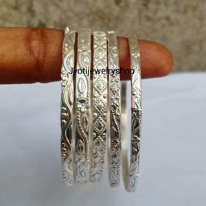 Op de afbeelding: Een set van vijf zilverkleurige metalen armbanden met verschillende reliëfpatronen, waaronder golvende lijnen, bloemmotieven en geometrische ruitvormen, naast elkaar geplaatst met ingewikkelde texturen en een gepolijste afwerking.