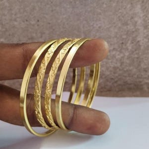 以下が含まれることがあります： ゴールドカラーの金属製バングル4本セット。2本は滑らかで光沢のある表面で、残りの2本には外周全体に複雑な花の模様が繰り返し刻印されています。シンプルで洗練されたデザインのアクセサリーです。