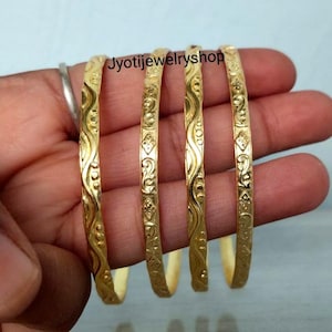 Può includere: Quattro braccialetti rigidi color oro con un motivo in rilievo ripetuto di linee ondulate e piccole forme geometriche. La superficie metallica ha una finitura lucida e il testo Jyotijewelryshop è visibile sopra i braccialetti.