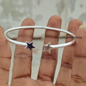 May include: A silver-toned open cuff bracelet featuring two five-pointed star charms at the ends of the band. The metal has a smooth, polished finish. The text Jyotijewelryshop is visible on the right side of the bracelet band.