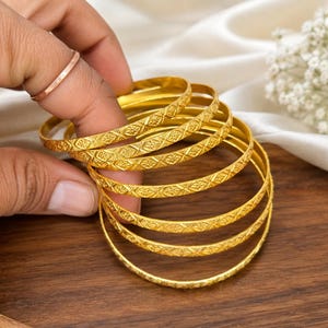 Op de afbeelding: Een set van zes dunne gouden armbanden, elk met een herhalend reliëfpatroon van ruiten en bloemen op het buitenoppervlak. De ronde armbanden zijn op elkaar gestapeld en vertonen een consistente metallic gele afwerking en textuur.