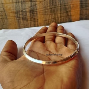 Puede incluir: Un brazalete rígido de metal plateado, liso y pulido, con un perfil redondeado y ligeramente cónico. La superficie del brazalete es reflectante y sencilla, con el texto Jyotijewelryshop impreso en pequeñas letras negras sobre el metal.