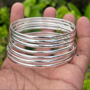 Peut inclure: Un ensemble de sept joncs fins de couleur argentée empilés ensemble. Chaque bracelet présente une surface lisse et arrondie avec une finition métallique réfléchissante. Les bracelets sont de forme circulaire et ont une épaisseur uniforme.