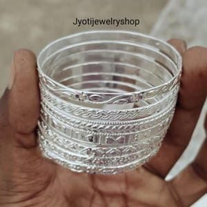 Puede incluir: Una pila de siete brazaletes de color plateado con varios patrones en relieve, incluyendo diseños florales, trenzados y geométricos. El texto Jyotijewelryshop es visible en la parte superior de la imagen. Cada brazalete tiene un acabado metálico pulido.