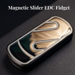 Op de afbeelding: Een rechthoekig magnetisch Magnetic Slider EDC Fidget speeltje met een metallic afwerking met goudkleurige en donkergroene wervelpatronen. Het apparaat heeft een glad, gepolijst oppervlak met een duidelijke rand en de tekst Magnetic Slider EDC Fidget.