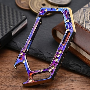 May include: A hexagonal metal carabiner featuring a vibrant, heat-anodized finish with iridescent shades of blue, purple, and gold. The frame includes a spring-loaded gate, a built-in bottle opener at the base, and decorative circular cutouts.