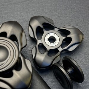 Könnte beinhalten: Zwei mattschwarze Fidget Spinner mit drei Flügeln, Aussparungen und einem silberfarbenen Kugellager in der Mitte. Einer ist von oben zu sehen, während der andere teilweise mit einer Seitenansicht seiner runden Kappe dargestellt ist.