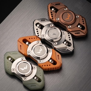 May include: Four fidget spinners arranged diagonally, featuring a central silver circular bearing with a stylised W logo. The spinners have outer frames in matte sage green, orange, brushed silver, and bronze, each with visible metal ball bearings.