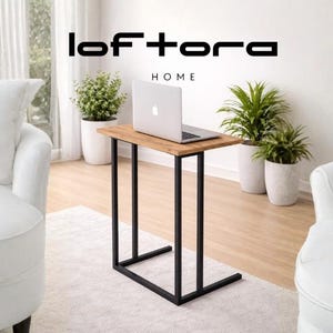 May include: A rectangular side table with a light brown wood-grain top and a black metal frame, positioned between two white rounded armchairs. The table features the text loftora HOME above a silver laptop placed on its surface.
