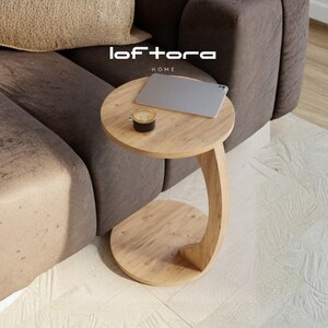 May include: A light brown wooden C-shaped side table with a circular top and base. The table features the text loftora HOME on the surface. A silver laptop and a small glass cup containing a dark liquid are placed on the table top.