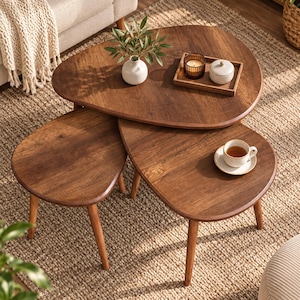 May include: A set of three nesting coffee tables with a dark wood finish and rounded, organic shapes. The tables feature tapered wooden legs and are arranged in a tiered formation. A small white vase with green leaves, a candle, and a cup sit on top.