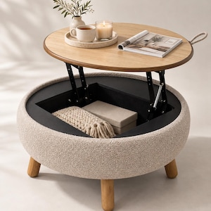 May include: A round coffee table with a light oak wood top and a cream-colored textured fabric base. The table features a lift-top mechanism revealing a black interior storage compartment containing a beige knit throw and a rectangular box.