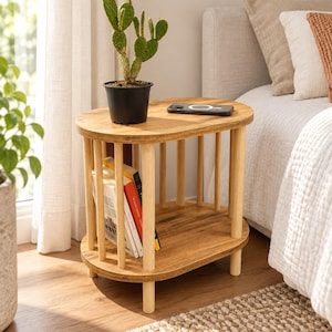 May include: An oval wooden side table with a slatted base design, featuring a smooth top surface and a lower shelf. The table is light brown with vertical cylindrical supports. A green cactus in a black pot and a black smartphone rest on the top.