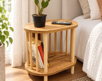 Puede incluir: Una mesa auxiliar ovalada de madera con diseño de base de listones, que cuenta con una superficie superior lisa y un estante inferior. La mesa es de color marrón claro con soportes cilíndricos verticales. Un cactus verde en una maceta negra y un smartphone negro descansan sobre ella.