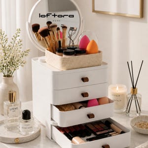 May include: A white multi-drawer makeup organizer with brown leather-like pulls, topped with a round mirror featuring the text loftora HOME. The unit holds makeup brushes, sponges, and cosmetics in drawers and a woven basket on the top surface.
