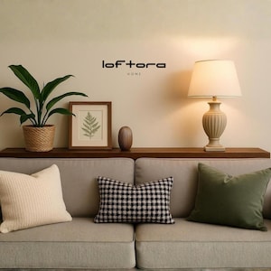 May include: A beige sofa with three throw pillows: one cream, one black and white checkered, and one olive green. Above the sofa, a wooden shelf holds a potted plant, a framed fern print, a small brown vase, and a lamp with the text loftora HOME.