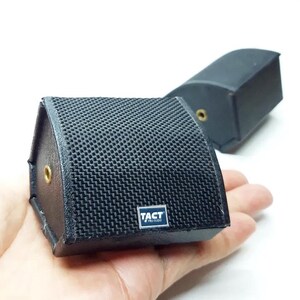 Pode incluir: Dois pequenos alto-falantes pretos em forma de cunha com uma grade frontal de malha texturizada. Cada alto-falante possui uma porta de entrada circular dourada na lateral e uma pequena placa de logotipo azul e branca com TACT PRO-AUDIO.