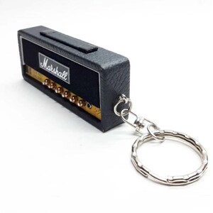 May include: A black rectangular keychain shaped like a miniature guitar amplifier. The front features the white Marshall logo on a black background with a gold-colored control panel containing several small knobs and a silver metal keyring attached.