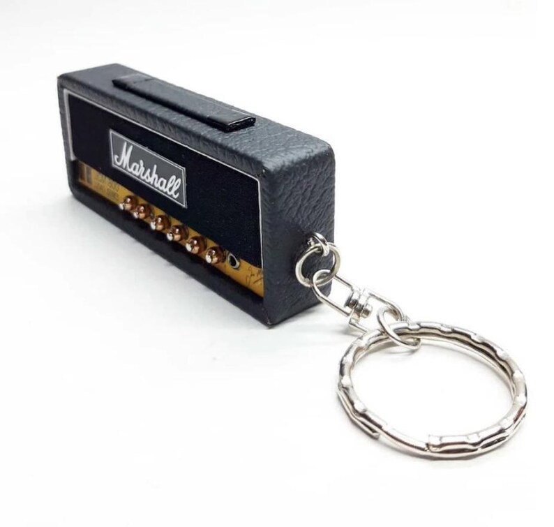May include: A black rectangular keychain shaped like a miniature guitar amplifier. The front features the white Marshall logo on a black background with a gold-colored control panel containing several small knobs and a silver metal keyring attached.