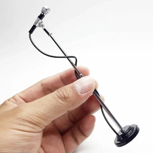 May include: A miniature microphone on a silver-coloured stand with a black circular base. The microphone head is silver and black, connected by a thin black cable that wraps around the straight metal stand, extending down to the base.