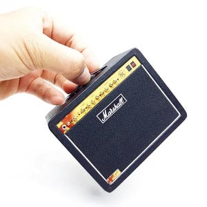 May include: A miniature black guitar amplifier with a textured finish, featuring a gold-colored control panel with knobs, a black speaker grille, and the white Marshall logo printed on the front. The device includes a small handle on the top side.