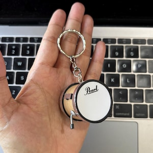 Könnte beinhalten: Ein Miniatur-Trommel-Schlüsselanhänger mit einem zylindrischen Holzkörper und einem weißen Trommelfell. Auf dem Trommelfell ist der schwarze Schriftzug Pearl aufgedruckt. Der Schlüsselanhänger hat einen silbernen Metallring.