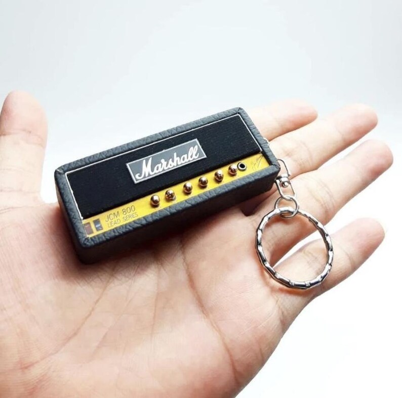May include: A miniature rectangular keychain modeled after a Marshall JCM-800 Lead Series guitar amplifier. The device features a black textured exterior, a yellow control panel with small gold knobs, and the white Marshall logo on the front.
