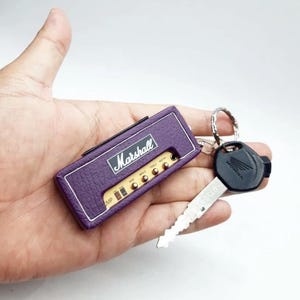 May include: A purple rectangular keychain designed to resemble a Marshall guitar amplifier, featuring the white Marshall logo, control knobs, and a silver metal ring attached to a black Honda motorcycle key with a silver metal blade.
