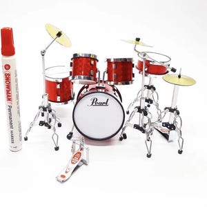 以下が含まれることがあります： Pearlのロゴが入ったバスドラム、複数のタム、銀色のスタンドに取り付けられた金属製のシンバルを備えた赤いミニチュアドラムセット。左側に置かれた白いSnowmanの油性マーカーは、セットの高さが約14cmであることを示しています。