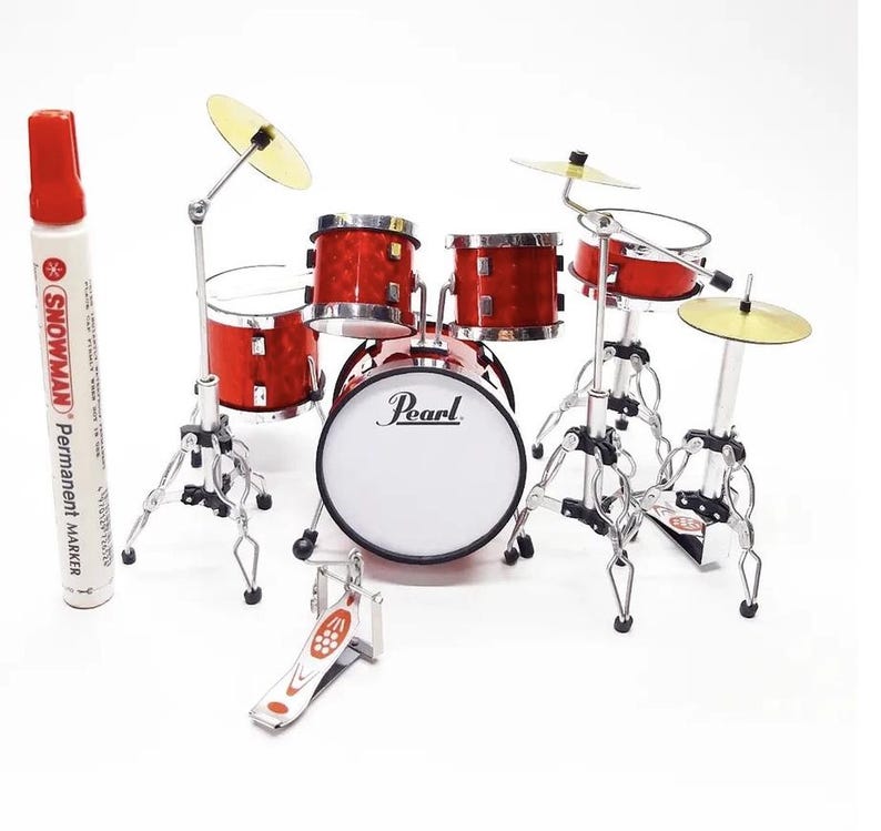 以下が含まれることがあります： Pearlのロゴが入ったバスドラム、複数のタム、銀色のスタンドに取り付けられた金属製のシンバルを備えた赤いミニチュアドラムセット。左側に置かれた白いSnowmanの油性マーカーは、セットの高さが約14cmであることを示しています。