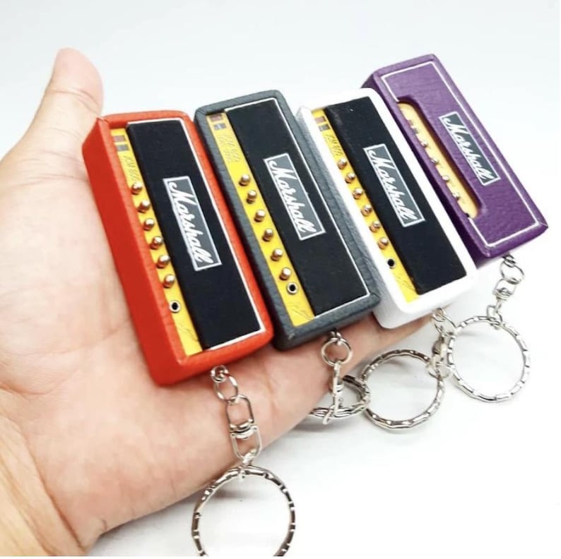 May include: Four miniature amplifier keychains in red, black, white, and purple. Each rectangular unit features a black front panel with the white Marshall logo, gold-colored control knobs, and a silver metal split ring attached to the base.
