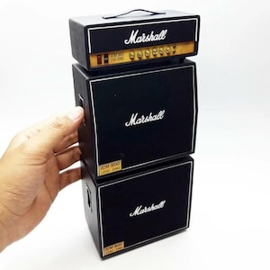 Op de afbeelding: Een miniatuur Marshall JCM 800 versterkerkop gestapeld op twee bijpassende luidsprekerkasten. De zwarte eenheden hebben witte Marshall-logo's, gouden bedieningspanelen met knoppen en kleine gouden labels met JCM 800 LEAD 1960.