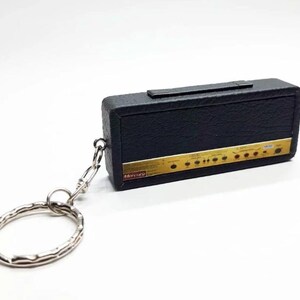 May include: A black rectangular keychain shaped like a guitar amplifier with a textured surface, a top handle, and a gold-colored control panel featuring knobs and the text Mercury. A silver metal split ring is attached by a small chain.
