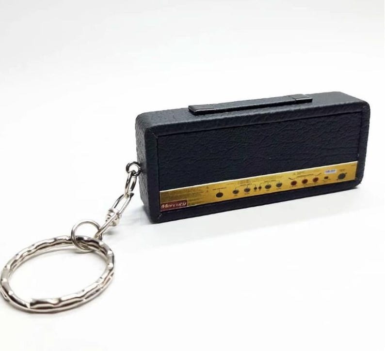 May include: A black rectangular keychain shaped like a guitar amplifier with a textured surface, a top handle, and a gold-colored control panel featuring knobs and the text Mercury. A silver metal split ring is attached by a small chain.