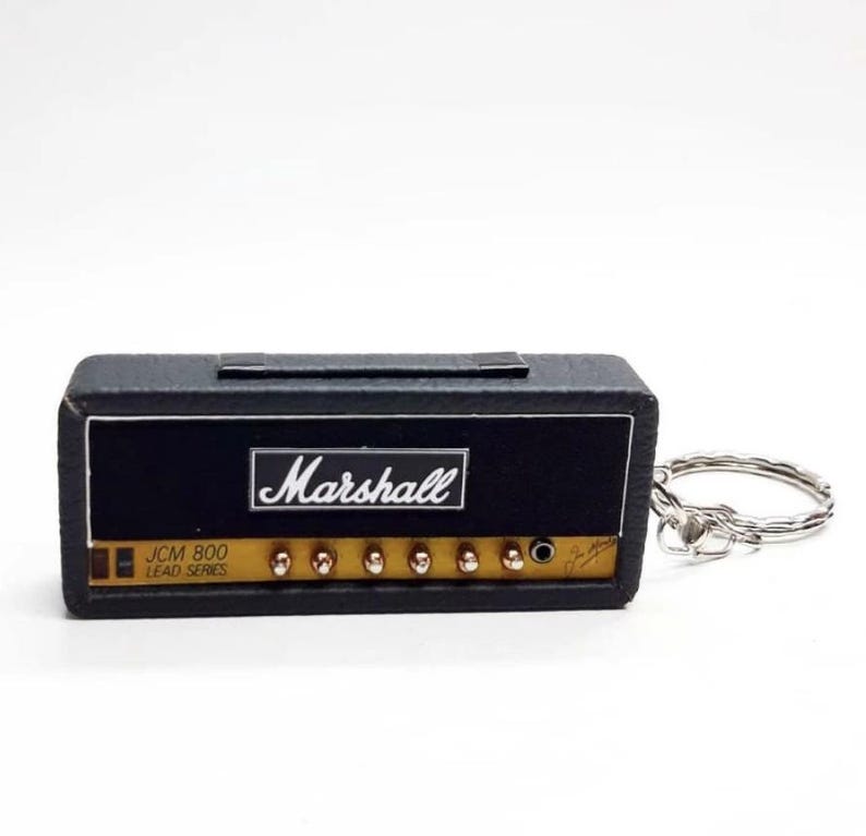 May include: A miniature black guitar amplifier keychain featuring the Marshall logo in white text. The front panel includes a gold-colored control section with knobs, a black input jack, and the text JCM 800 LEAD SERIES printed in black.