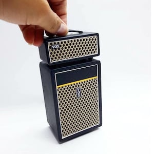 May include: A miniature Vox guitar amplifier stack featuring a black textured exterior, a beige honeycomb speaker grille, and a thin yellow horizontal stripe. The brand name VOX is printed in white on a black rectangular badge on both components.