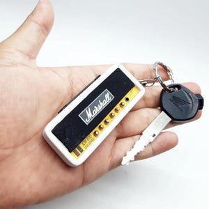 May include: A rectangular keychain designed to resemble a Marshall JCM 800 amplifier head, featuring a black front panel with white Marshall branding, gold-colored knobs, and a white casing, attached to a metal ring with a black Honda ignition key.
