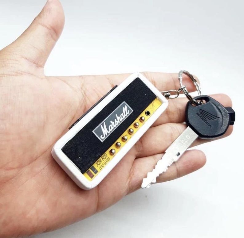 May include: A rectangular keychain designed to resemble a Marshall JCM 800 amplifier head, featuring a black front panel with white Marshall branding, gold-colored knobs, and a white casing, attached to a metal ring with a black Honda ignition key.