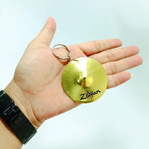 Puede incluir: Un llavero circular de metal dorado con forma de platillo, que presenta una cúpula central elevada y el texto negro Zildjian impreso en la superficie. Esta réplica de instrumento musical está unida a una anilla de metal plateado.