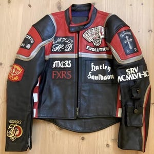 May include: A black and red leather motorcycle jacket featuring various patches including Harley Davidson, Sturgis 50th anniversary, and a skull playing card design. The jacket has a front zip, long sleeves with small utility pockets, and white accents.