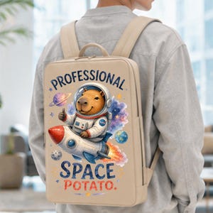 May include: A beige backpack featuring a graphic of a capybara in a space suit riding a rocket ship. The text reads PROFESSIONAL SPACE POTATO. The design includes a planet, stars, and a nebula in blue, purple, and orange colors against the beige fabric.