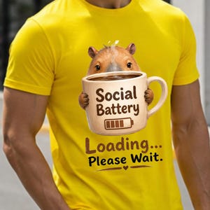 May include: A bright yellow short-sleeved t-shirt featuring a graphic of a capybara holding a beige coffee mug. The mug displays the text Social Battery with a battery icon, and below it, the text Loading... Please Wait. with a small heart icon.