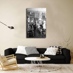 K&ouml;nnte beinhalten: Ein schwarz-wei&szlig;es Wandkunst-Poster, das eine Vintage-Pilates-Studio-Szene zeigt. Das Bild zeigt Personen, die h&ouml;lzerne Trainingsger&auml;te mit Federn und Riemen benutzen, w&auml;hrend gerahmte Bilder und ein gro&szlig;er Spiegel die Studiow&auml;nde schm&uuml;cken.