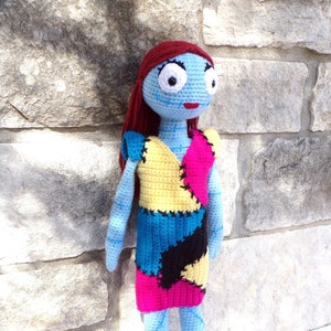 May include: A crocheted doll with blue skin, red hair, and a colourful patchwork dress. The doll has large, white eyes and a red mouth. The doll is standing on a white surface in front of a stone wall.
