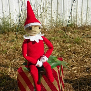 May include: A red and white striped gift box with a green ribbon. A crocheted elf doll wearing a red hat and white collar is sitting on top of the box.