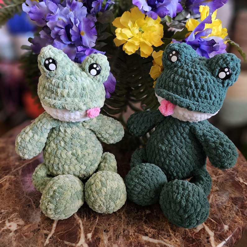 Puede incluir: Dos peluches de rana hechos a mano con ganchillo en tonos verdes. Uno es verde claro, el otro es verde oscuro. Ambos tienen vientres blancos, lenguas rosas y ojos grandes y redondos. Est&aacute;n sentados sobre una superficie de m&aacute;rmol marr&oacute;n, con un fondo de flores moradas y amarillas.