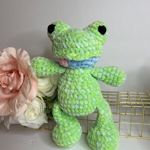 Puede incluir: Una rana de peluche hecha a mano en tonos verdes, azules y rosas. La rana tiene grandes ojos negros y una expresi&oacute;n sonriente. El material suave y texturizado le da a la rana una apariencia tierna. Es un art&iacute;culo artesanal caprichoso.